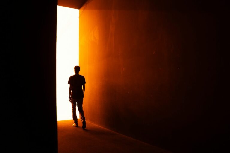 A person walks from a dark space toward a bright doorway, symbolizing letting go of old defenses and choosing growth.
