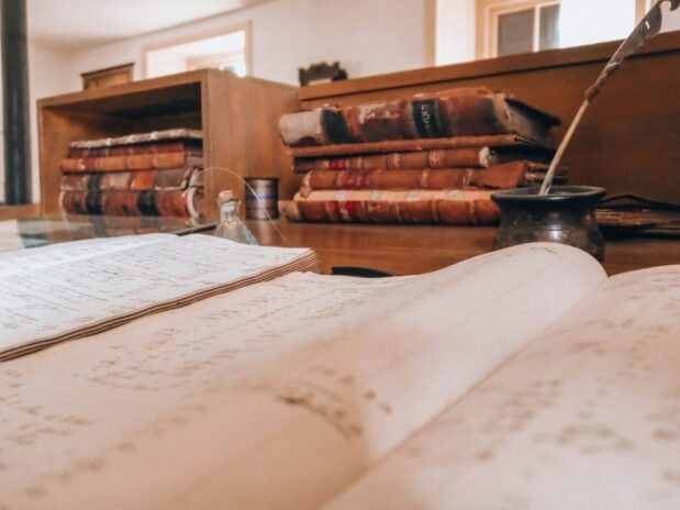 Open book, old books, and a quill arranged on a desk, representing reading, learning, and imagination